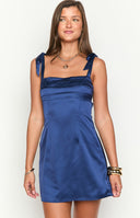 Model smiling in a navy blue satin mini dress- perfect for homecoming. The dress features a straight- pleated neckline and wide shoulder straps that tie into bows.