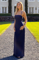 Navy blue satin maxi dress with thin spaghetti straps- a square neckline- and a low back with criss-cross detailing. A light blue bracelet is worn.