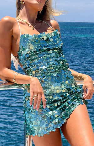 a woman leans a against a railing wearing an aqua sequin mini dress and gold bracelets with the ocean in the background