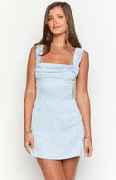 brunette model wearing a blue floral mini dress with gold pendant necklace standing at a bar