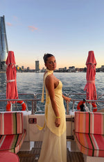 Side view of a yellow satin maxi dress with a scarf halter neck, open back with long ties, and a sheer overlay skirt