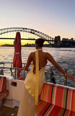 Rear view of a yellow satin halter neck maxi dress with an open back, thin criss-cross straps, and a wide vertical scarf deta