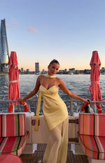 Yellow satin cowl neck maxi dress with thin straps, a long draped scarf, and a sheer panel skirt with a high slit