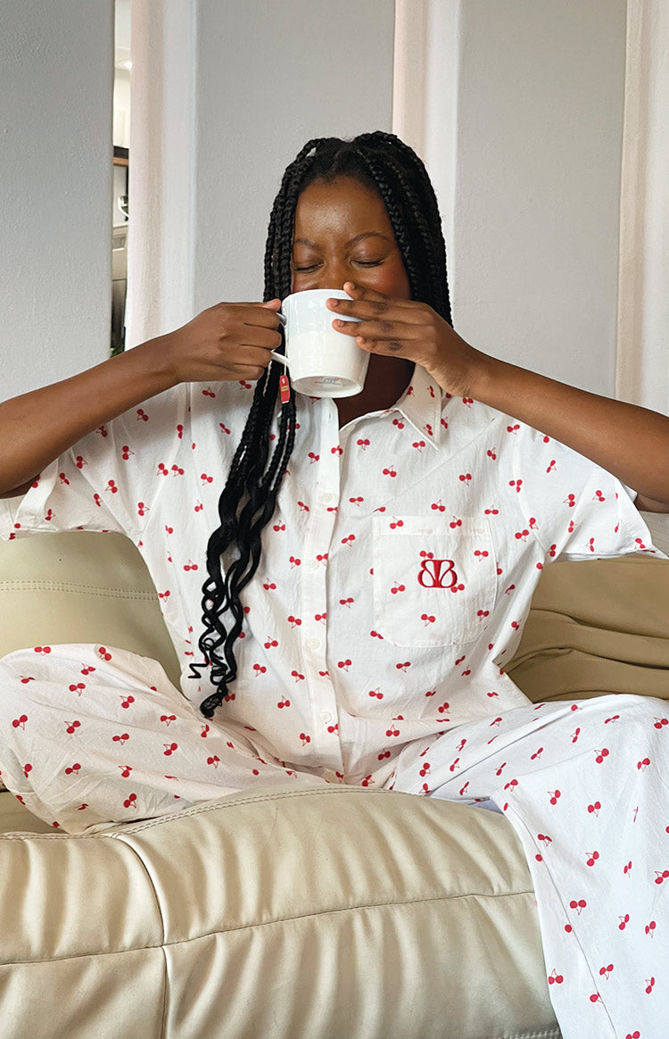 White cherry print pajama shirt with a red embroidered logo on the chest pocket and matching printed pants