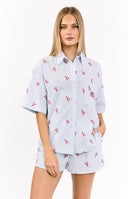 A woman in a light blue and white striped pajama shirt with a red lobster print- unbuttoned over a white ribbed tank top- and matching blue and white striped shorts with the same red lobster print.