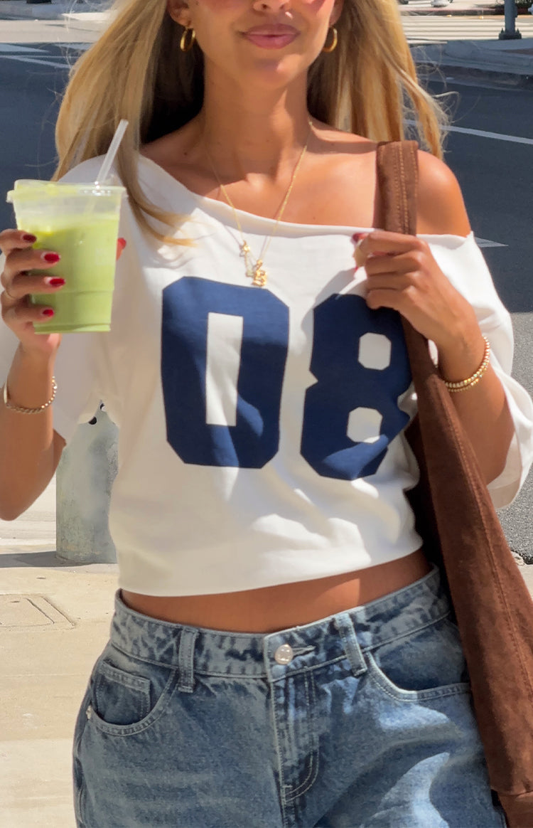 A close-up of a white crop top with a wide off-the-shoulder neckline featuring a large navy blue 08 graphic on the chest.