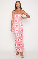 A person with blonde hair and sunglasses models a white strapless maxi dress with a red geometric pattern. They are wearing gold jewelry and carrying a small black handbag while standing in a sunlit outdoor corridor.