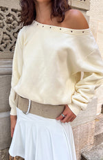 Cream off-shoulder sweater with an asymmetric neckline featuring silver eyelets and long ribbed cuffs