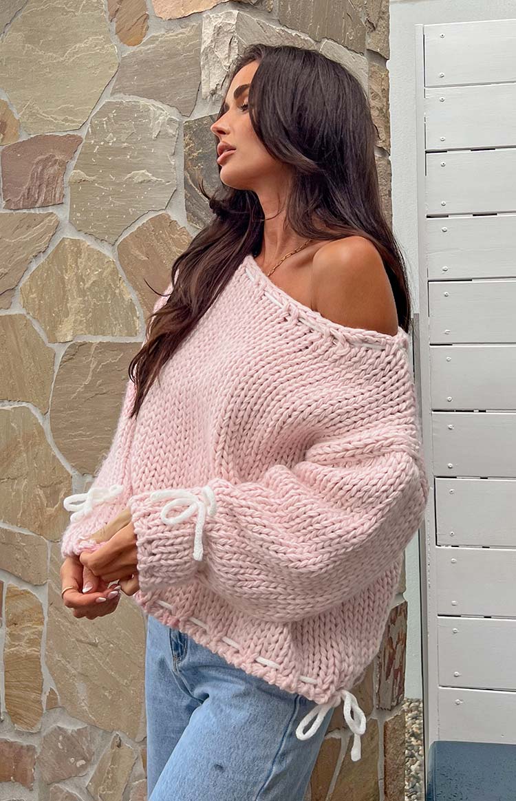 Rear view of a baby pink chunky knit sweater with white stitching details and ties on the shoulders and cuffs
