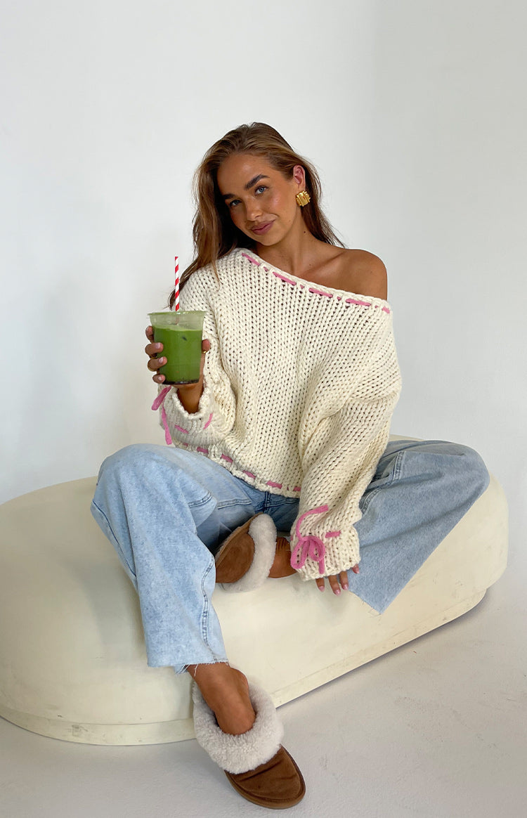 Cream chunky knit sweater with off-shoulder neckline, pink stitching, and bow details on the cuffs
