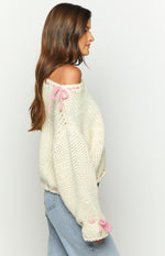Side view of a cream chunky knit sweater with pink bow ties on the off-shoulder neckline and ruffled cuffs