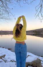 Rear view of a cropped yellow chunky knit sweater with white tie details on the sleeves and a curved hem