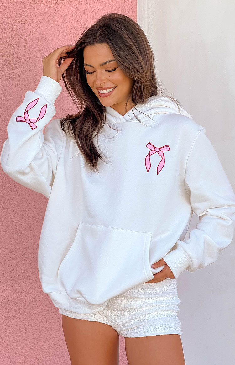 Brunette girl wearing a big oversized white hoodie with pink bow designs embroidered on the chest and sleeves. Girl is wearing white ruffled shorts.