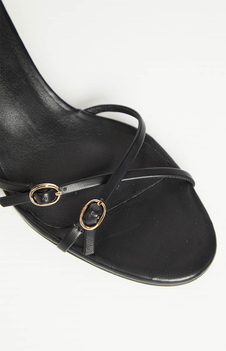 Close-up of the black heels showing thin front straps with two small gold buckles and a square toe