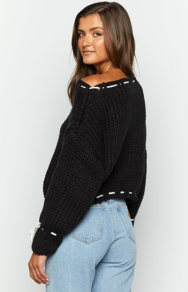 Close-up of the black chunky knit sweater showing white tie detail on the cuff and white stitching along the hem