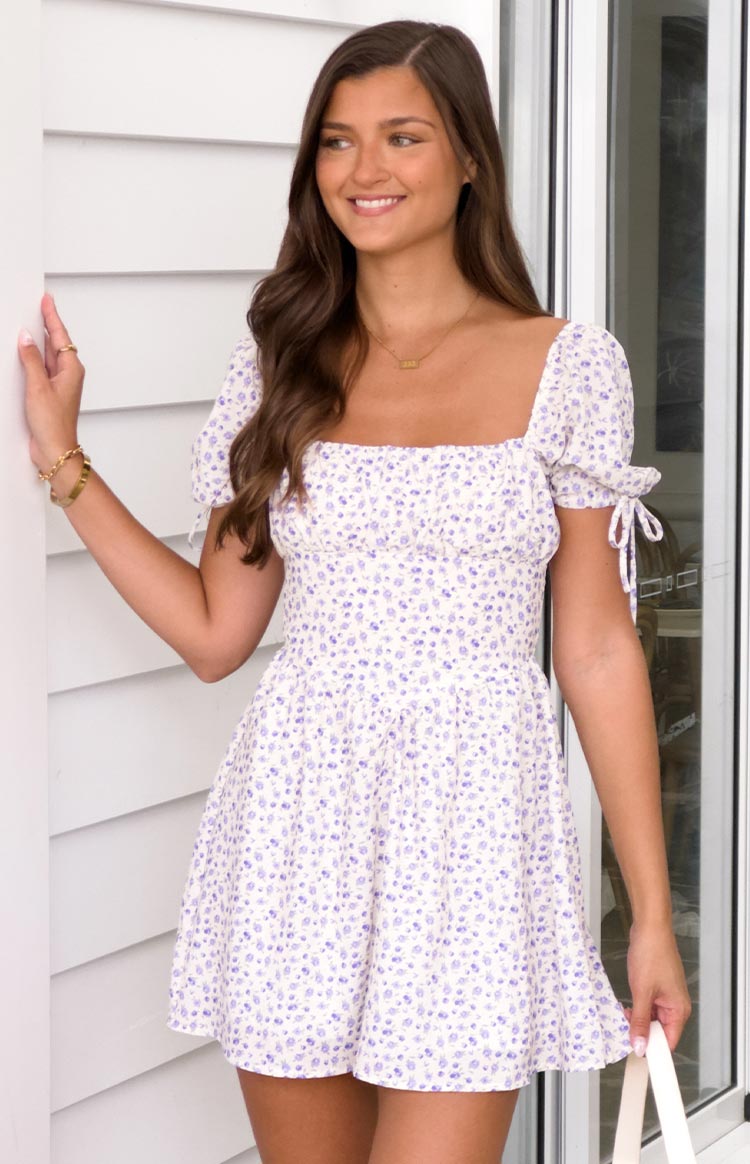 Front view of a white mini dress with purple floral print, square neckline, puff sleeves with tie details, and a smocked bodice