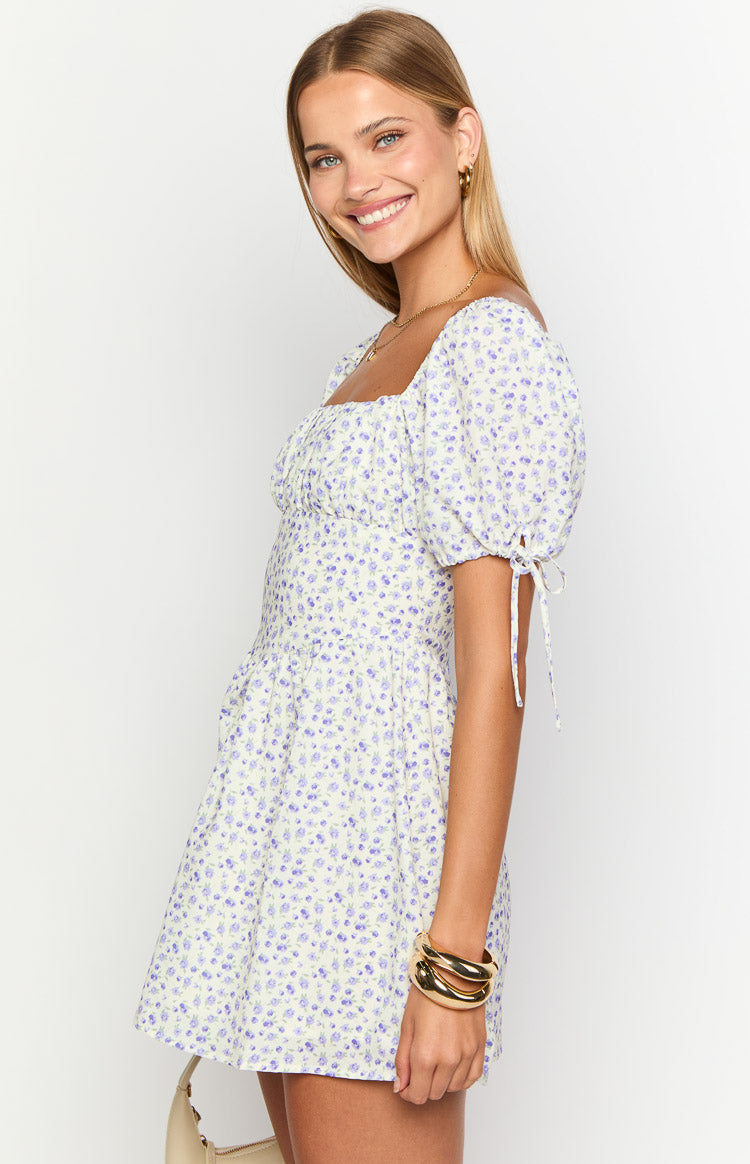 Side view of a light yellow mini dress with purple floral print, square neckline, ruched bust, and puff sleeves with tie cuffs