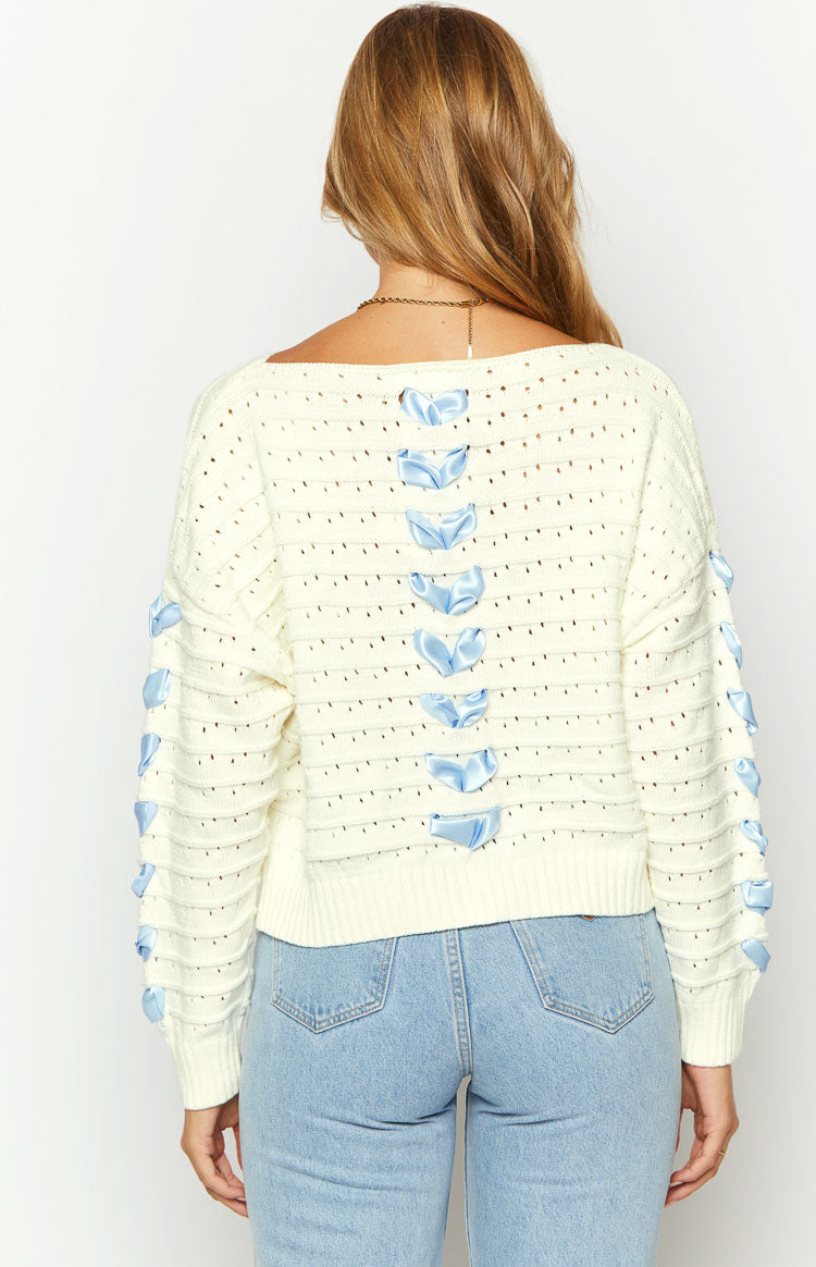 Front view of a cream knit sweater with blue ribbon bows on the chest and long sleeves, featuring a boat neck and ribbed cuffs