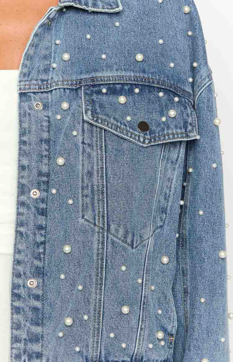 Close-up of a mid-wash denim jacket's chest pocket with flap and button, featuring scattered pearl embellishments