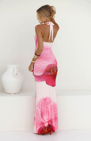 A back view of our model wearing a pale pink halter-style maxi dress with a large vibrant pink orchid print. The dress has a low-cut back with a tie at the neck perfect for your garden party.
