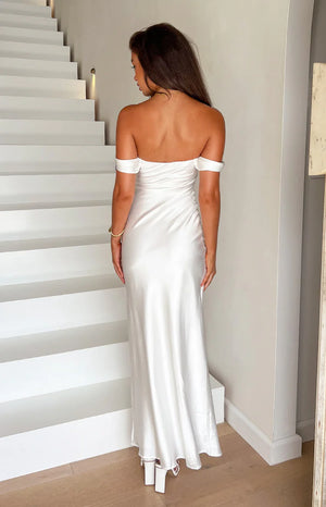 Our model wearing a floor length maxi dress in a shimmering white satin fabric. The dress features an off the shoulder neckline with folded sleeves and a subtle ruching detail along the center back seam. The smooth fabric is fitted and drapes elegantly down the back.