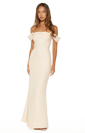 A full-length shot of our model wearing a form-fitting off-the-shoulder maxi dress in a light cream color. This dress features a draped wrap-style bodice and a long sleek skirt that pools slightly at the feet.