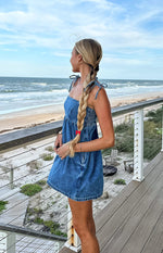 Side view of a blue denim mini dress featuring tie straps, a smocked bust, and a relaxed A-line skirt