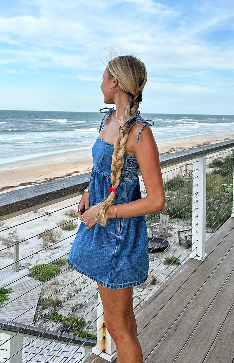 Side view of a blue denim mini dress featuring tie straps, a smocked bust, and a relaxed A-line skirt