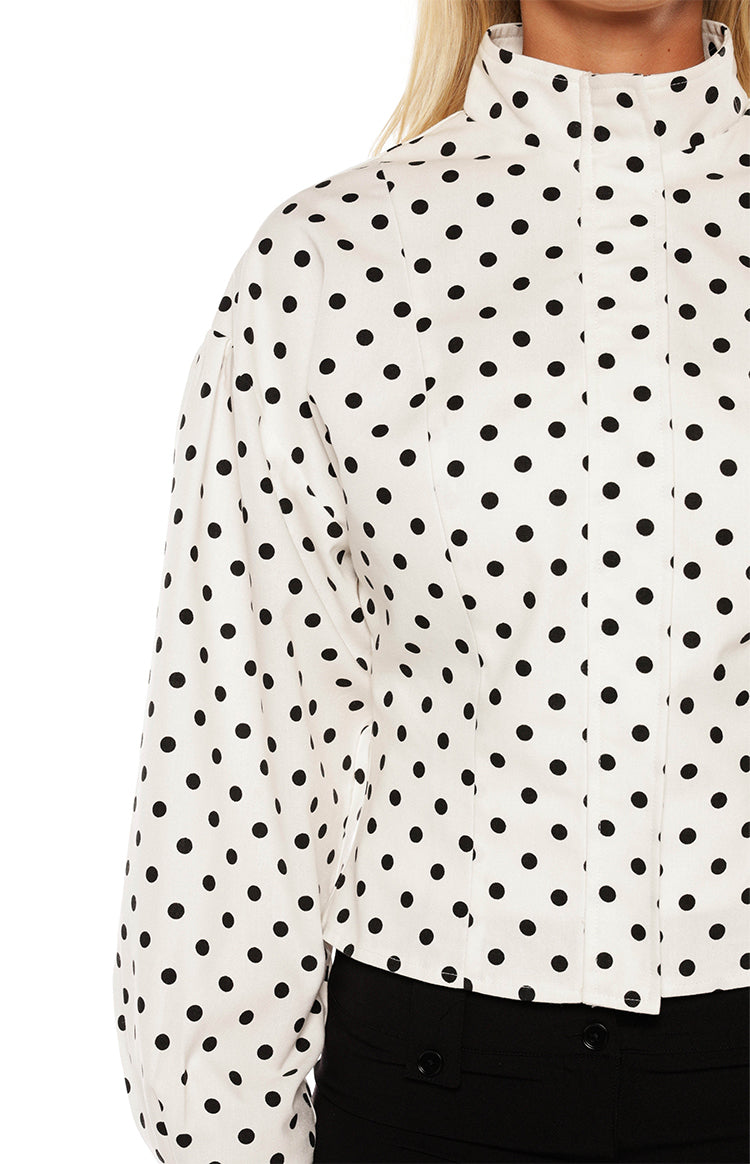  A close-up of a white and black polka dot top featuring a mock neck collar a hidden placket and a puffed long sleeve.