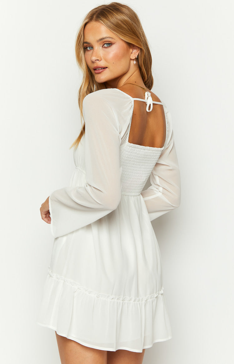 Side-rear view of a white chiffon mini dress with a square back, shirred panel, tie detail, sheer long sleeves, and ruffled hem