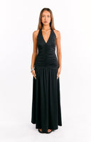 A female model wearing a black halter maxi dress with a plunging V-neckline and a ruched bodice. The skirt is loose and flowing and the model is also wearing black heeled sandals.