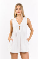 A woman stands outdoors in a sleeveless white playsuit featuring a V-neck with a tie at the bust and vertical pintuck detailing. She is wearing tall- matte green rain boots over white socks that peek out at the top.