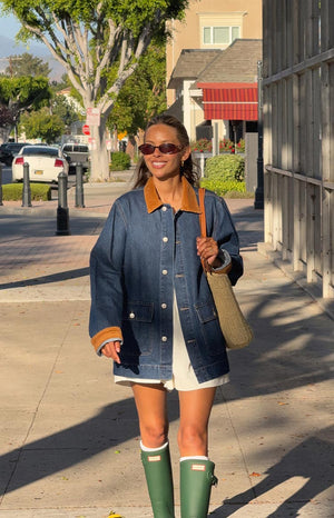 A woman walks on a sidewalk wearing an oversized- mid-wash denim jacket with a contrasting tan corduroy collar and corduroy trim on the cuffs and pocket flaps. A white outfit is visible underneath- paired with tall green rain boots and white socks.