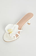 A close-up shows a person's feet wearing white heeled sandals adorned with white and yellow orchid accents. A gold ankle bracelet is visible on one ankle