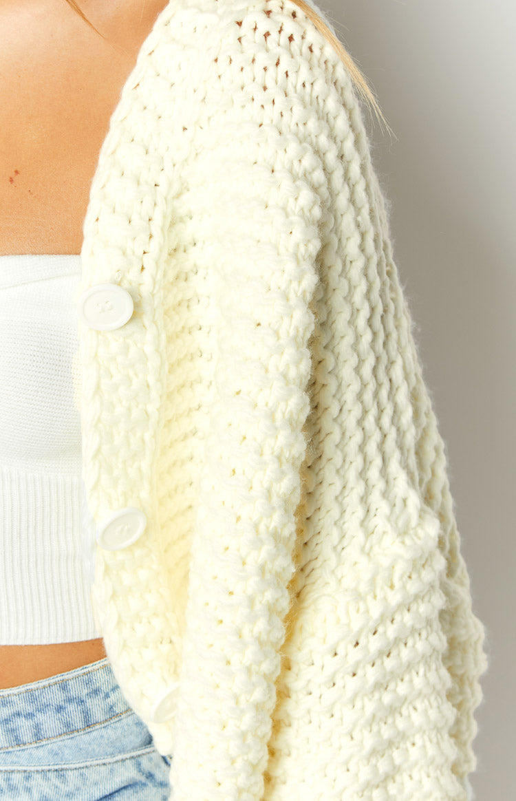 Close-up of the textured cream knit cardigan showing the cable knit pattern, white buttons, and side pocket detail