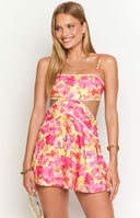 Brunette girl is wearing a mini dress with a cut out design and bright floral print. The dress has white trim details and girl is holding a white shoulder bag.