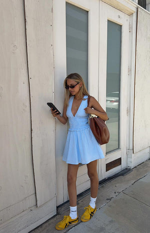 A woman wears a sleeveless mini dress in a light blue and white pinstripe fabric. The dress is designed with a V-neck- a shirred waist- and an A-line skirt- and is paired with white socks and bright yellow sneakers.