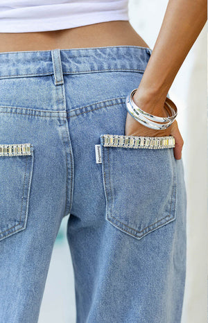 A close-up view of the back of a pair of light-wash denim jeans. The standout feature is the crystal embellishment—two horizontal rows of rectangular clear baguette-cut rhinestones are set along the top edge of each back patch pocket. The jeans have a straight or wide-leg cut and belt loops.