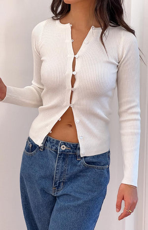 A white ribbed knit button-up long-sleeve top- partially unbuttoned to reveal the midriff- paired with mid-wash blue jeans for a casual look.