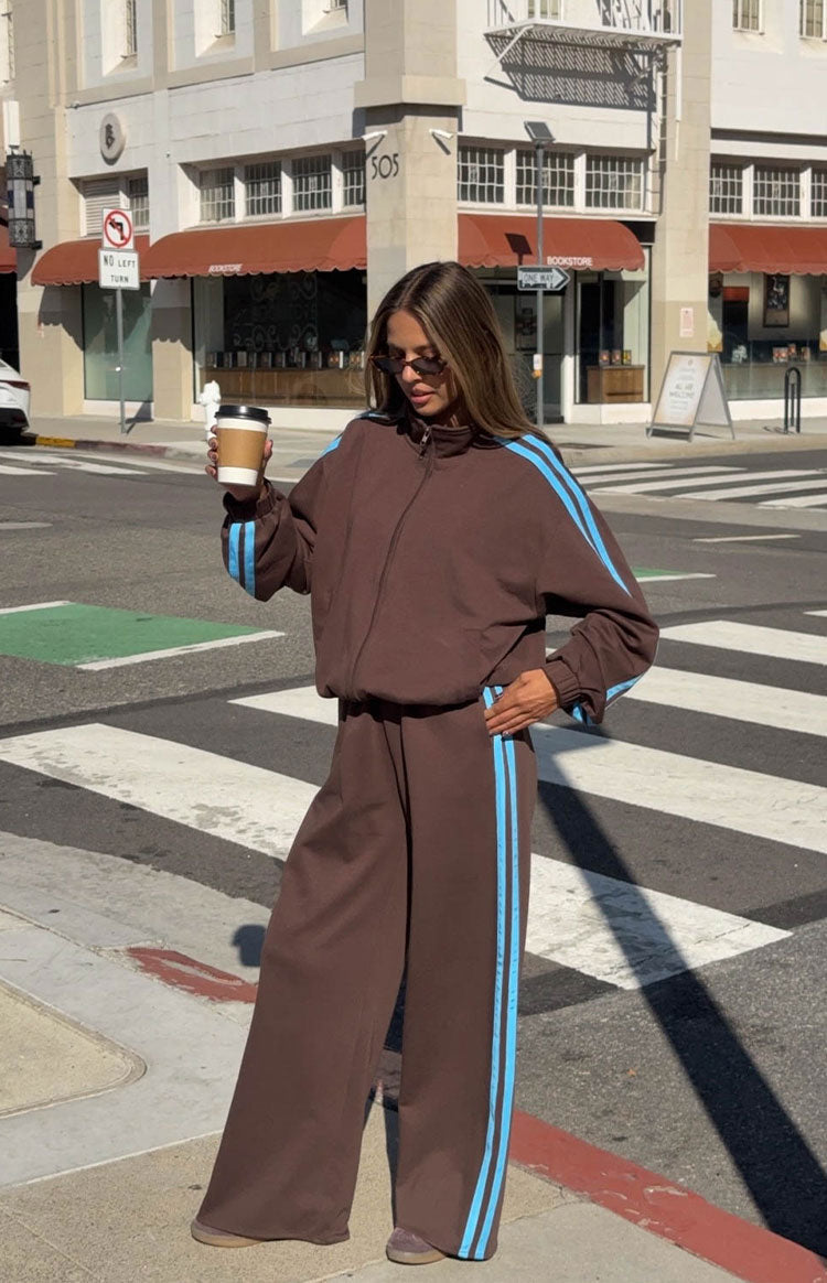 Full view of a chocolate brown zip-up jacket with blue stripes on the sleeves and matching wide-leg pants with blue side stripes