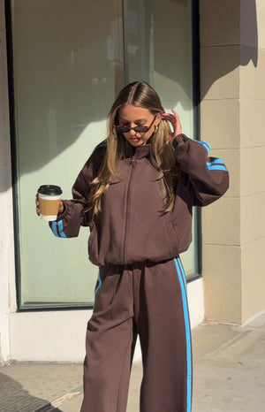 A female model wearing an oversized brown zip-up track jacket with a high collar and light blue stripes down the arms.