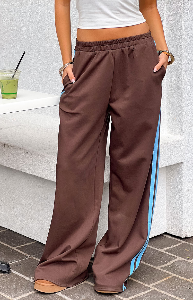 Front view of chocolate brown track pants with elasticated waist, side pockets, and light blue side stripe