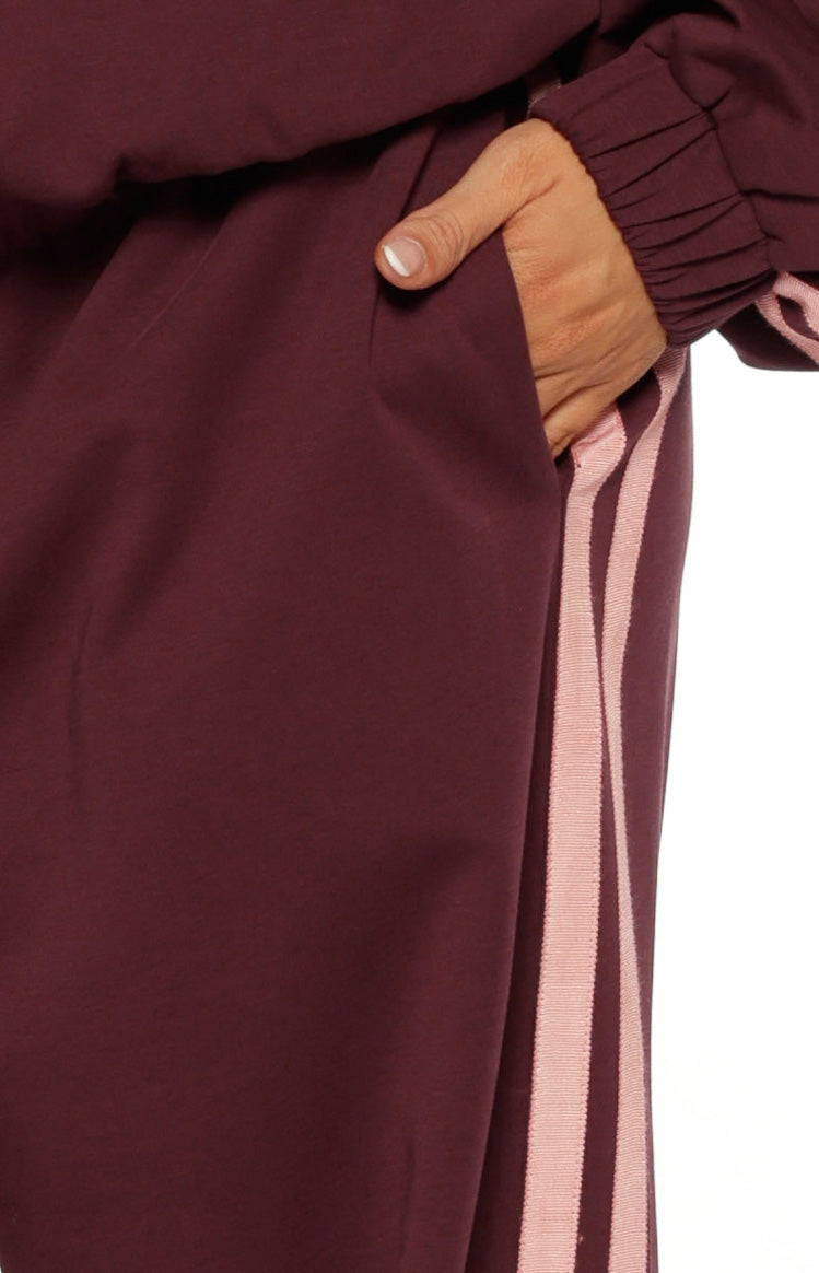 Detail shot of burgundy track pants showing the side pocket opening and vertical pink stripe