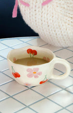 Angled view of a cream ceramic mug with wavy rim, red cherry and pink floral pattern, showing the handle and interior