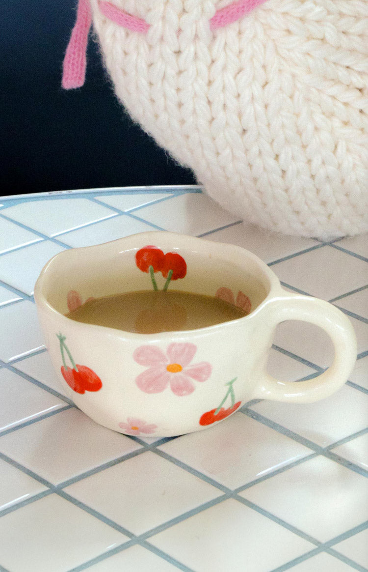 Angled view of a cream ceramic mug with wavy rim, red cherry and pink floral pattern, showing the handle and interior