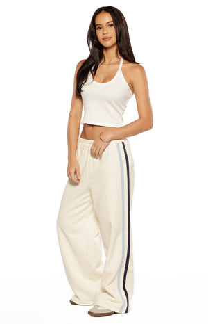 A lifestyle shot of our model standing on a tennis court wearing cream-colored fleece-style track pants. These pants feature a wide straight-leg silhouette and a thick elasticized waistband.