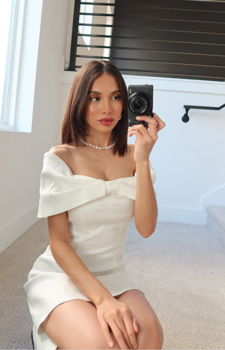 Close-up of the white off-shoulder mini dress showing the large bow detail at the bust
