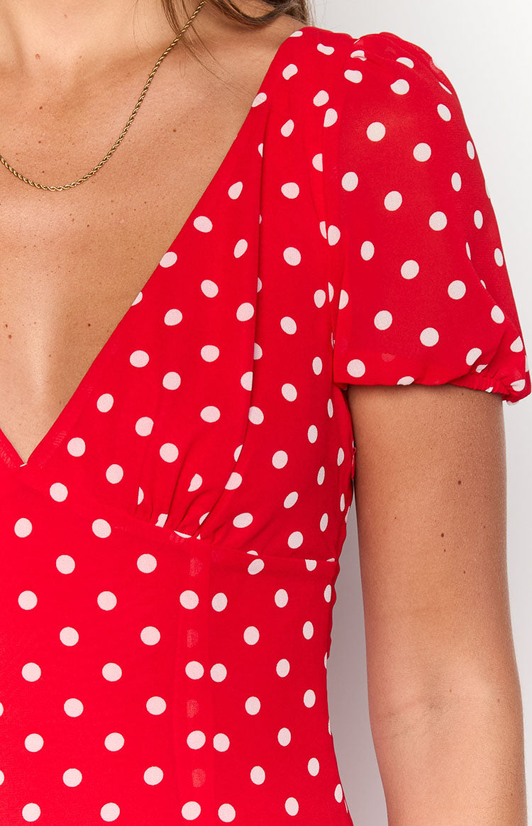 A close-up of a maxi dress in a vibrant red fabric with an all-over print of small white polka dots. This maxi features a deep V-neckline a fitted and gathered bust and a short slightly puffed sleeve with elasticized gathering at the cuff.