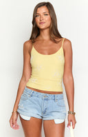 Close up of girl wearing a yellow cami with embroidered flower detailing and a white ruffle mini skirt.