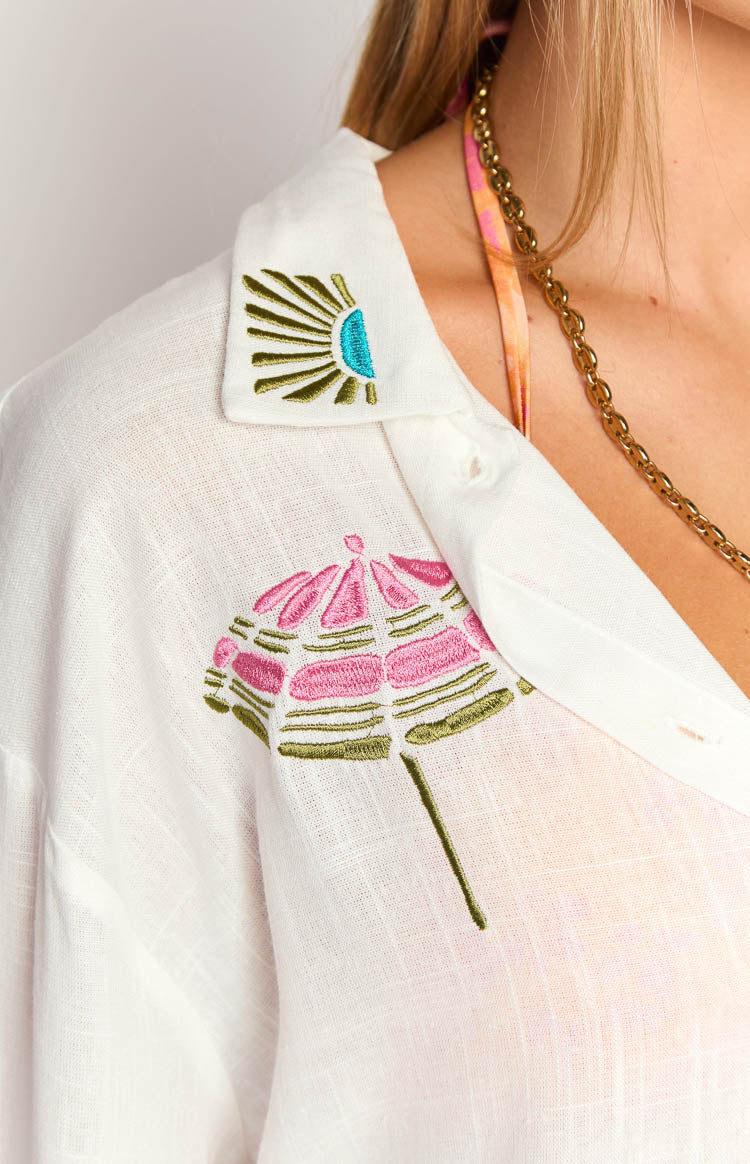 Close-up of the white long sleeve shirt with teal and olive sun embroidery on collar and pink beach umbrella on chest